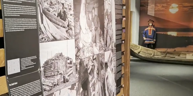 Mostra museale con pannelli informativi, foto storiche in bianco e nero e un manichino in abiti tradizionali Sami con barca d