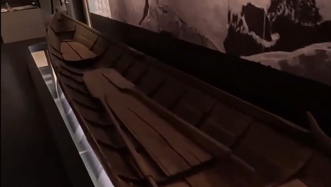Barca di legno tradizionale esposta in un museo, con remi all'interno. La barca è illuminata, con uno sfondo scuro e dettagli