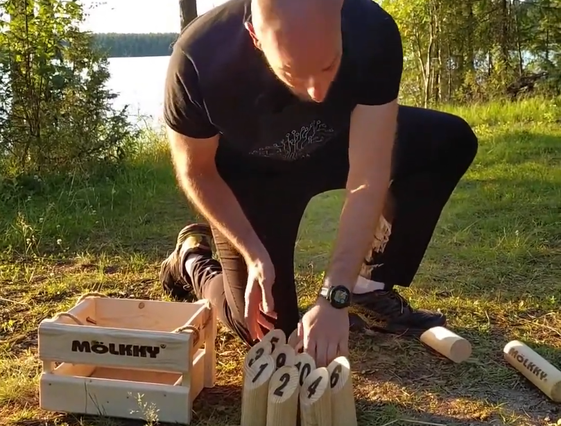Stefano Mini prepara il gioco Mölkky all'aperto, sistemando i birilli numerati vicino alla cassa di legno, con un lago sullo