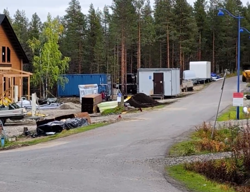 Cantiere con casa in costruzione, container blu, cabina portatile e materiali edili, circondato da alberi in una zona boschiv