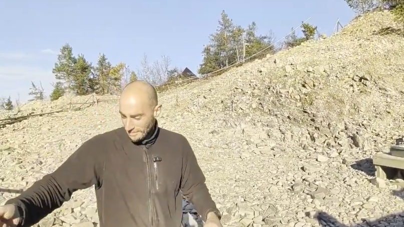 Stefano Mini in piedi su un terreno roccioso con alberi e una collina sullo sfondo, sotto un cielo azzurro e soleggiato.
