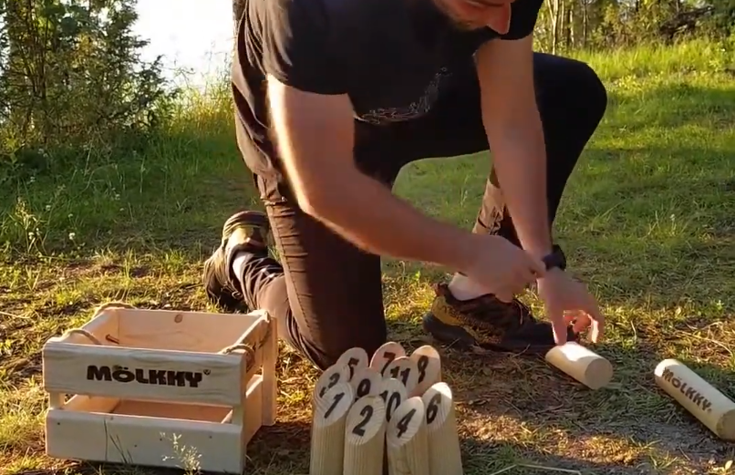 Stefano Mini si inginocchia sull'erba per giocare a Mölkky, con i birilli numerati e la cassa di legno visibili.