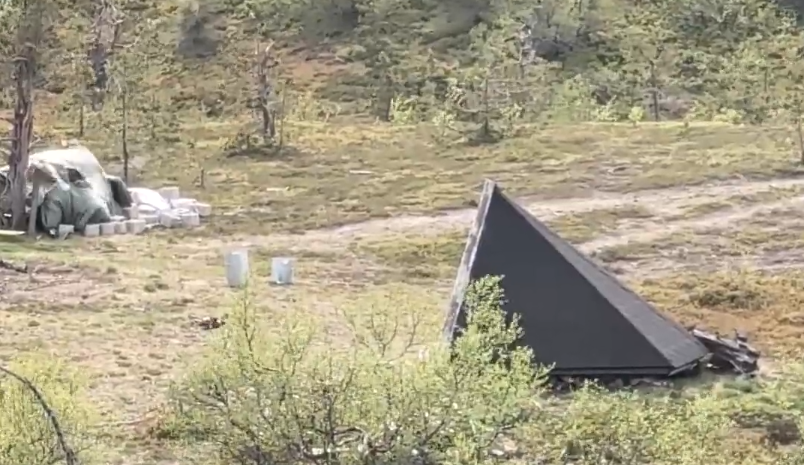 Capanna triangolare scura in un paesaggio rurale con alberi e materiali da costruzione coperti da un telo verde.