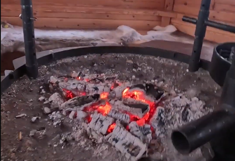 Braci ardenti e cenere in un braciere circolare, con pareti in legno e neve all'esterno. Atmosfera calda e accogliente.