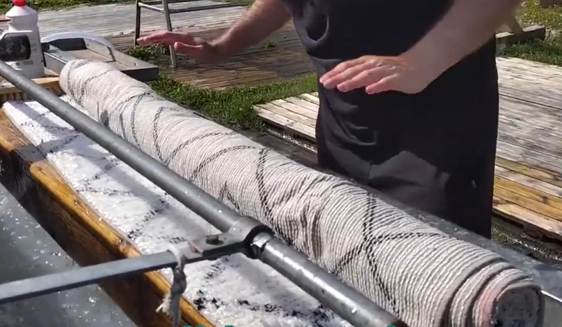 Stefano Mini lava un tappeto arrotolato con acqua e sapone, creando schiuma, su una superficie di legno all'aperto.