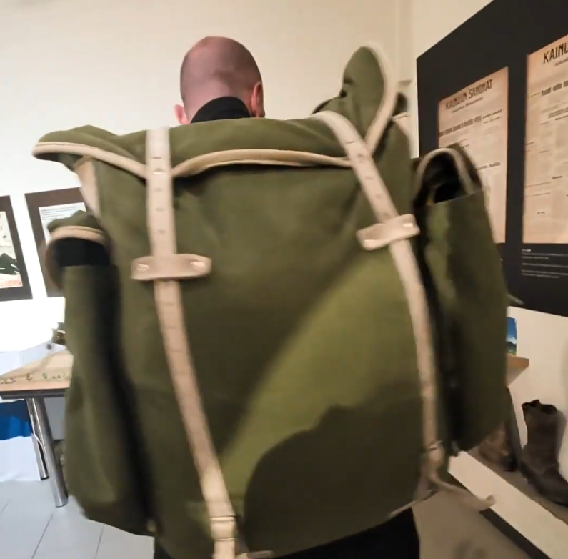 Stefano Mini di spalle con un grande zaino militare verde con cinghie chiare, in un ambiente che sembra un museo o una mostra