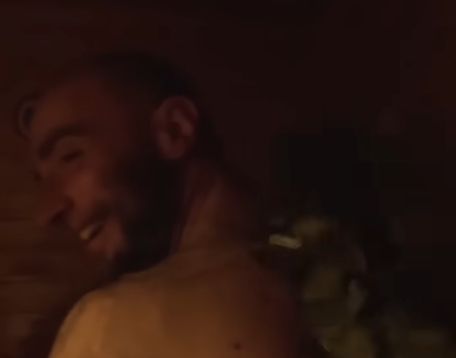 Stefano Mini sorride mentre si rilassa in una sauna, con un fascio di foglie per il massaggio sulla spalla.