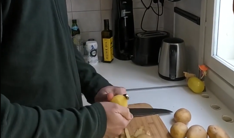 Stefano Mini pela una patata su un tagliere di legno in una cucina moderna, con altre patate, olio d'oliva e elettrodomestici