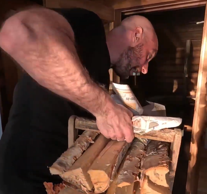 Stefano Mini si china per sistemare la legna da ardere e l'accendifuoco in un ambiente interno con pareti in legno.