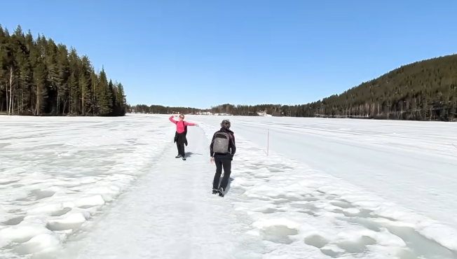 Due persone camminano su un lago ghiacciato e innevato sotto un cielo azzurro. Una donna in rosa saluta, Stefano Mini con zai