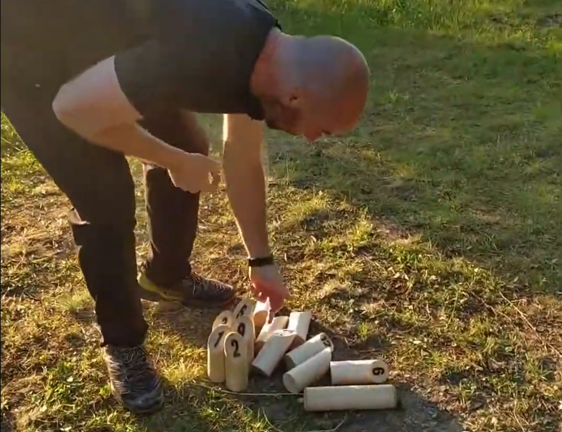 Stefano Mini si china per raccogliere o sistemare i birilli di legno numerati del gioco Mölkky su un prato.