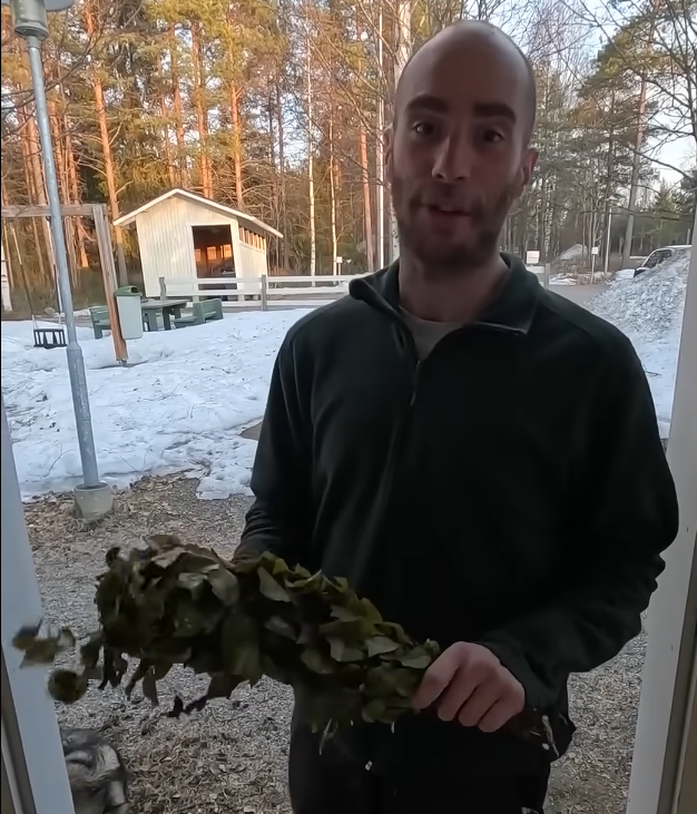 Stefano Mini tiene un fascio di foglie di betulla (vihta) per sauna, all'aperto in un paesaggio invernale con neve e alberi.