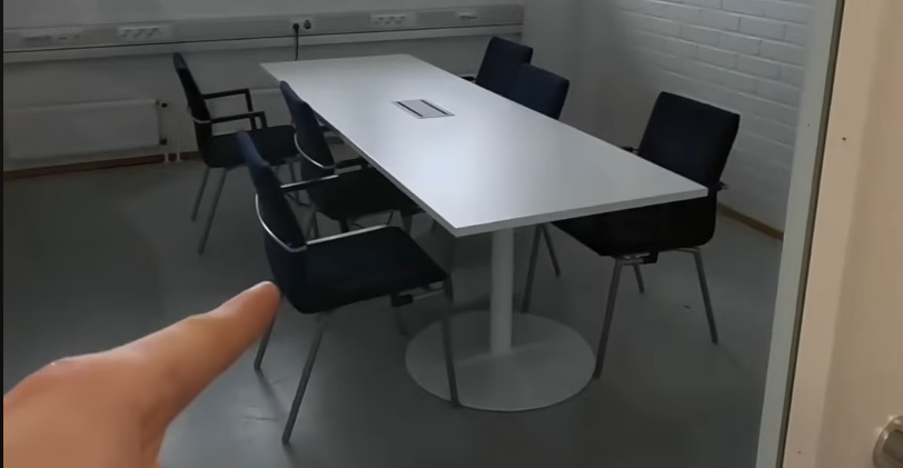 Sala riunioni moderna e vuota con tavolo bianco e sedie scure, una mano indica l'interno della stanza.