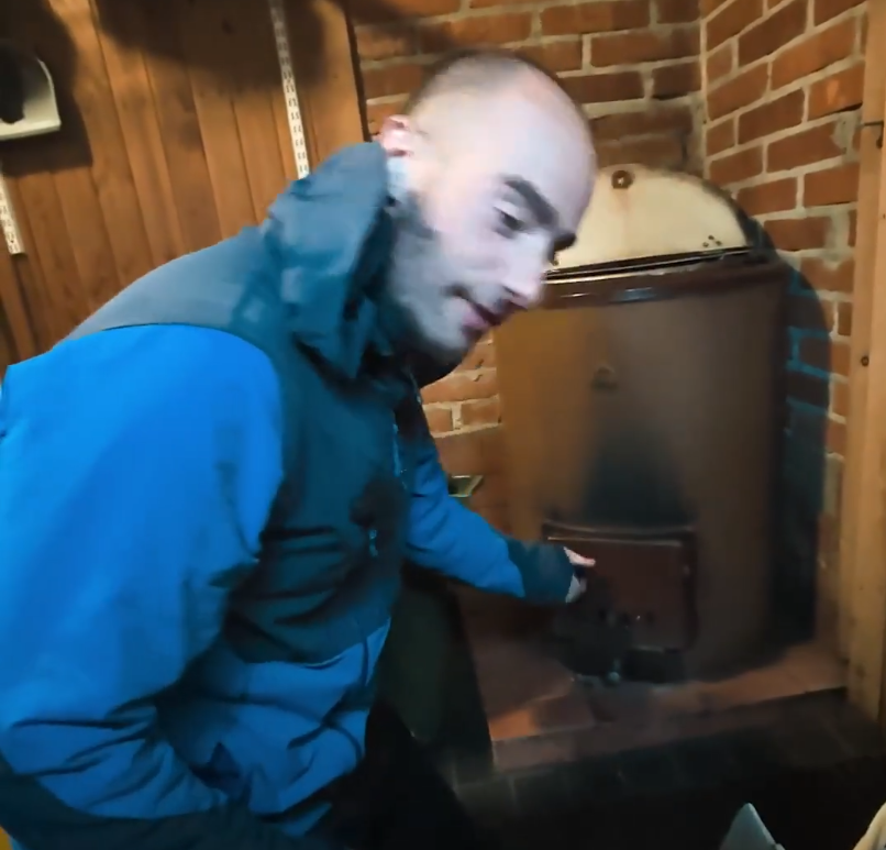 Stefano Mini in una stanza rustica con pareti in legno e mattoni, mentre apre lo sportello di una vecchia stufa a legna.