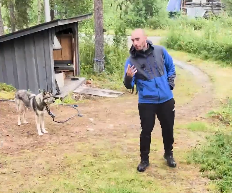 Stefano Mini con un cane husky legato vicino a una capanna di legno in un ambiente naturale e boschivo.