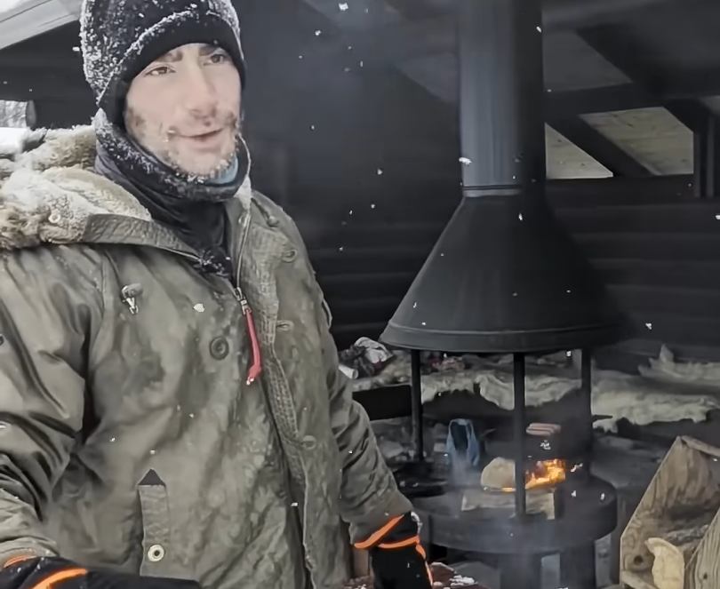 Stefano Mini in un rifugio innevato, indossa un cappello e una giacca invernale, mentre del cibo cuoce su un fuoco all'aperto