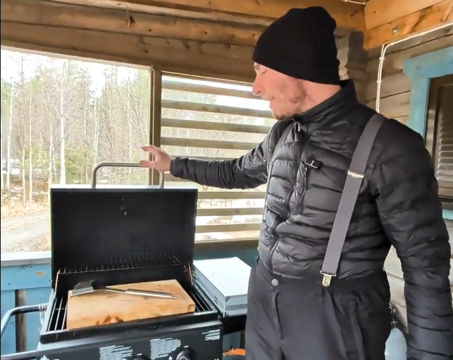 Stefano Mini apre un barbecue a gas su un portico, con una tavola di legno e una spatola all'interno, sullo sfondo alberi.