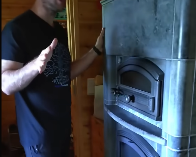 Stefano Mini gesticola accanto a una stufa a legna in maiolica grigia con porta in metallo e finestra.