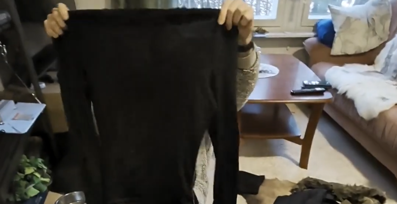 Mani che tengono in alto una maglia nera a maniche lunghe in un ambiente domestico.