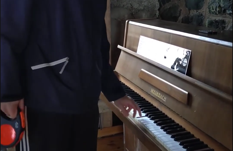 Stefano Mini suona un pianoforte verticale Weinbach, con spartito sul leggio e un guinzaglio per cani in mano.