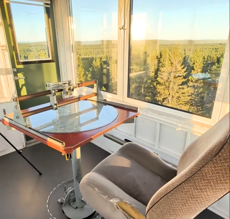 Interno di una torre di avvistamento con scrivania e sedia, vista panoramica su una vasta foresta di conifere al tramonto.
