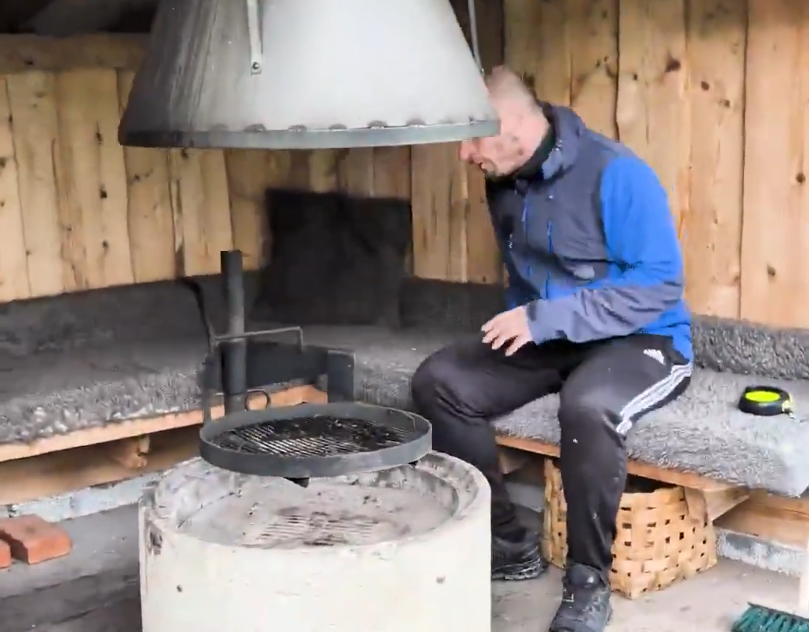 Stefano Mini si siede su una panca in una capanna di legno con un braciere e una griglia al centro, pronto per cucinare.