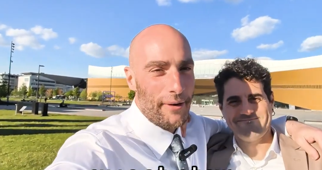 Stefano Mini e un amico sorridono in un selfie all'aperto, con un moderno edificio arancione e cielo blu sullo sfondo.