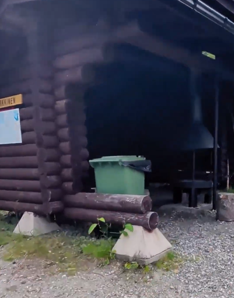 Capanna di tronchi in legno con un bidone verde all'esterno, su un terreno ghiaioso. Sembra un rifugio o un'area barbecue.