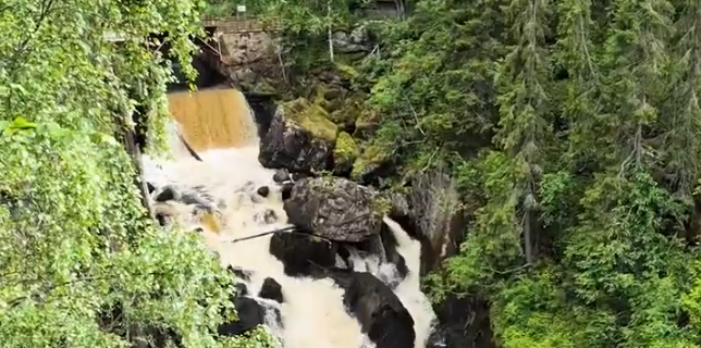 Cascata con acqua marrone che scorre impetuosa tra rocce e fitta vegetazione verde, con una struttura in legno in lontananza.