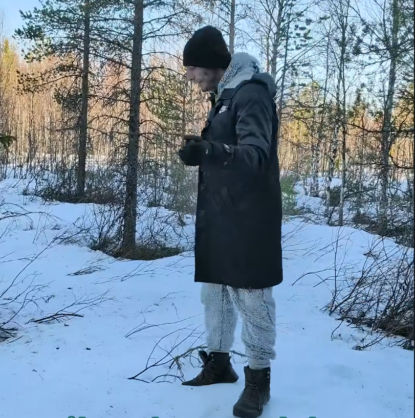 Stefano Mini in un bosco innevato, indossa un cappotto scuro e pantaloni chiari, guarda le sue mani. Giornata di sole.