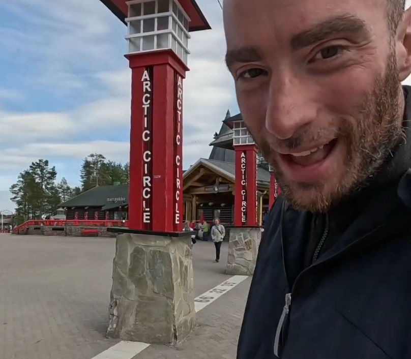 Stefano Mini sorride davanti ai pilastri rossi che segnano il Circolo Polare Artico, con edifici turistici sullo sfondo.