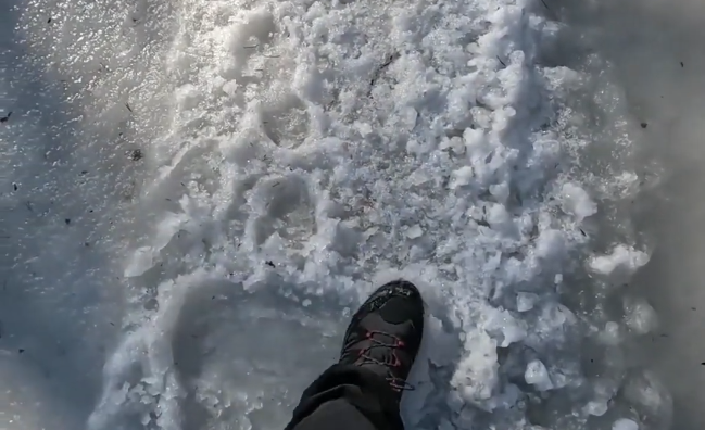 Piede con scarponcino da trekking che cammina su un sentiero innevato e ghiacciato, con frammenti di ghiaccio e neve sciolta.