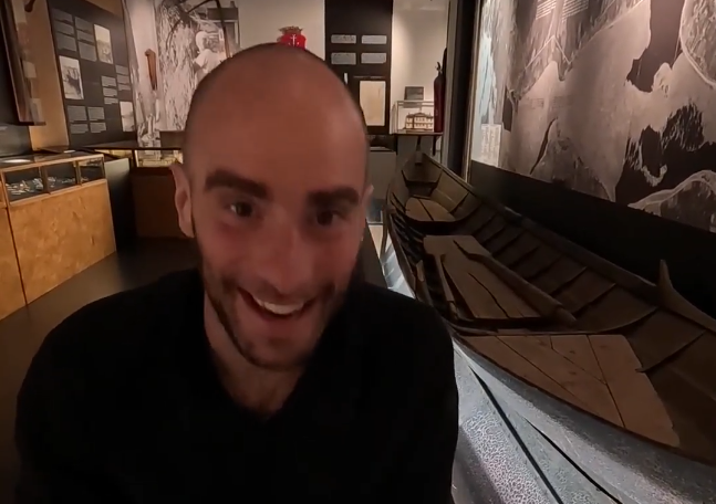 Stefano Mini sorride in un museo, con una barca di legno e mostre storiche sullo sfondo.