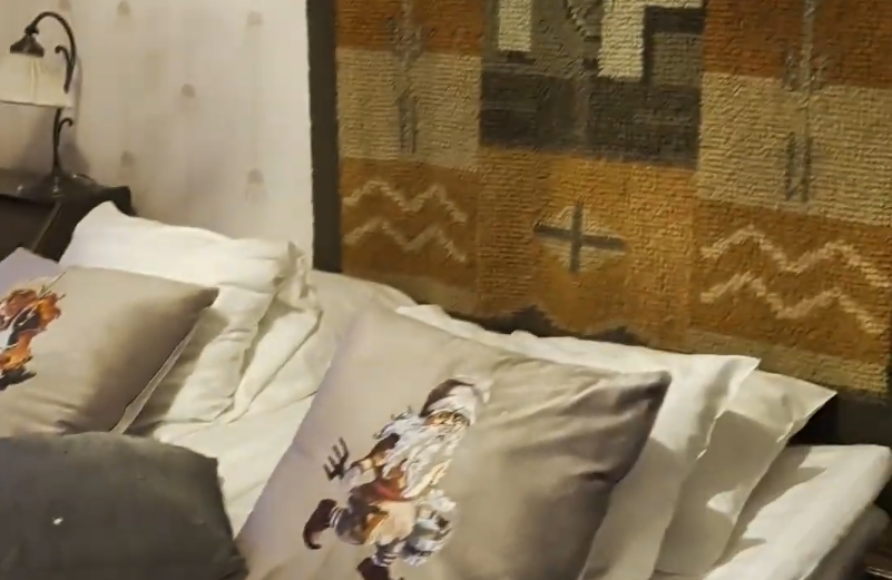 Letto con lenzuola bianche, cuscini decorati con gnomi, arazzo geometrico come testiera e lampada da comodino.