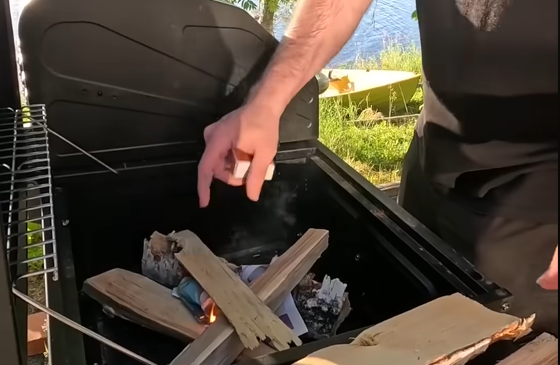 Stefano Mini accende il fuoco nel barbecue con legna e carta, preparando la griglia all'aperto vicino all'acqua.