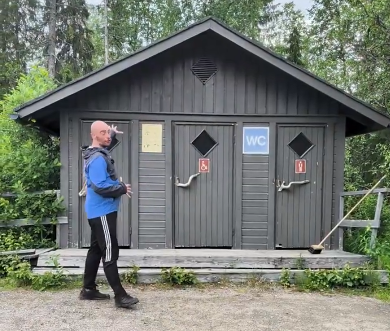 Stefano Mini in piedi davanti a un bagno pubblico in legno grigio con tre porte, una con simbolo WC e una per disabili.