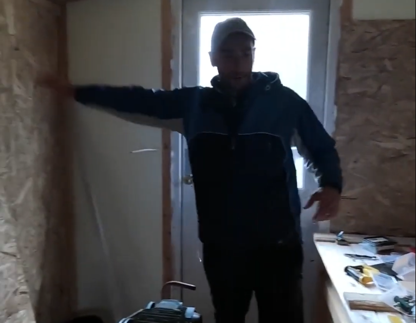 Stefano Mini in un ambiente di lavoro con pareti in OSB, indossa un cappellino e una giacca, con il braccio sinistro esteso.