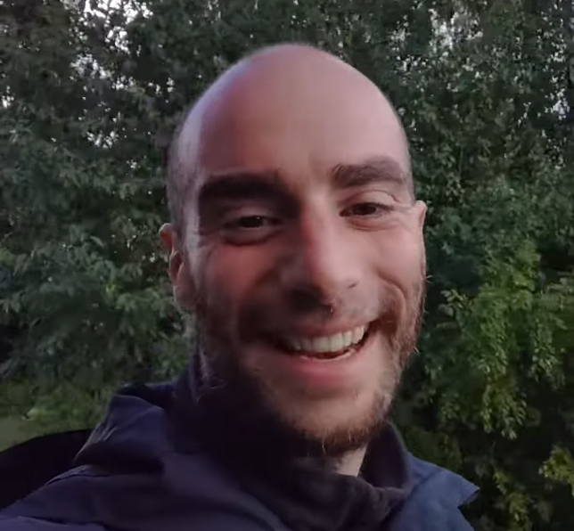 Stefano Mini, un uomo calvo con la barba, sorride felicemente alla telecamera con uno sfondo di alberi verdi.