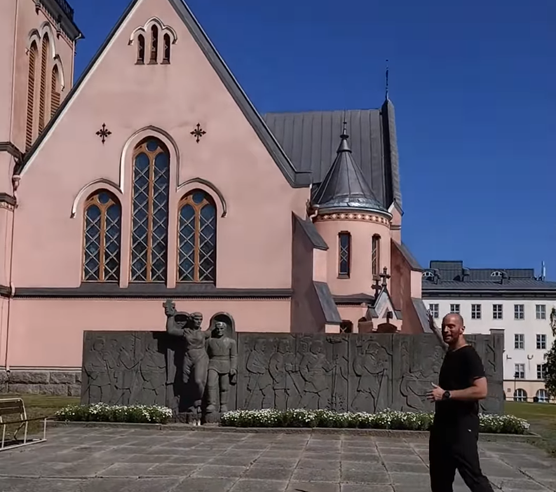 Stefano Mini in piedi davanti a una chiesa rosa con un monumento in pietra e un cielo azzurro.