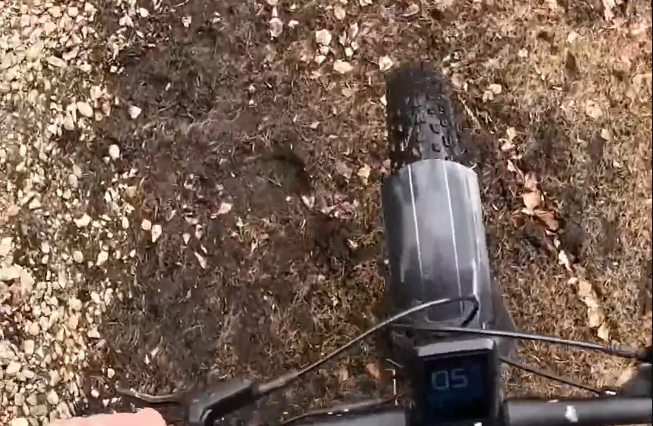 Vista in prima persona di una fat bike su un sentiero sterrato con ghiaia e foglie. Si vedono la ruota anteriore e il manubri