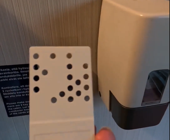 Una mano tiene un dispositivo bianco con fori, sullo sfondo un dispenser a muro e un cartello con istruzioni in più lingue.