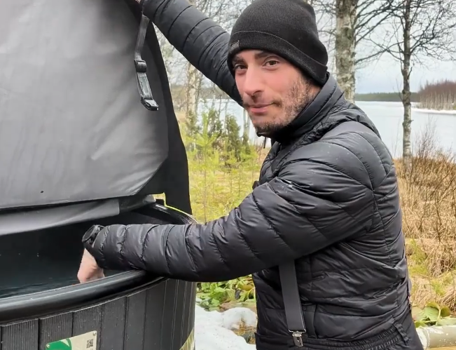 Stefano Mini apre il coperchio di una vasca idromassaggio esterna in un paesaggio invernale con lago ghiacciato sullo sfondo.