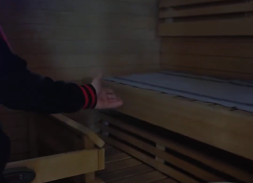 Il braccio di Stefano Mini gesticola all'interno di una sauna in legno con panche. L'ambiente è illuminato in modo soffuso.