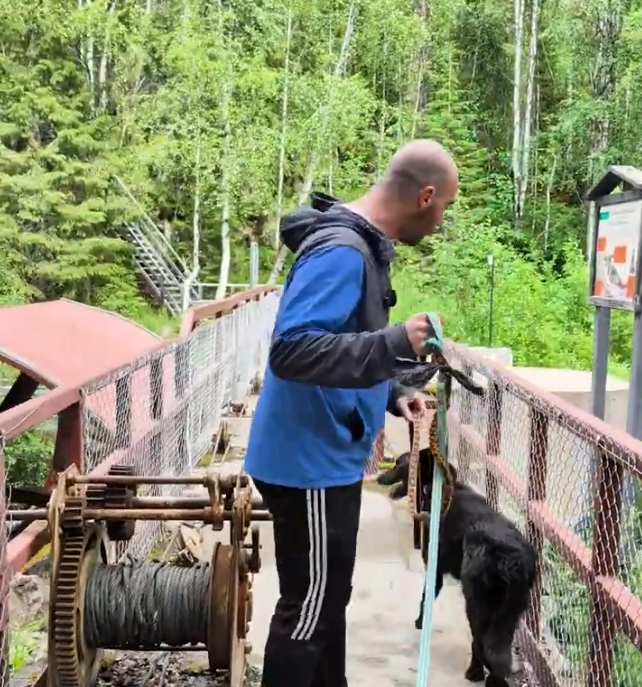 Stefano Mini e il suo cane nero su un ponte metallico con un argano arrugginito, circondati da alberi verdi.
