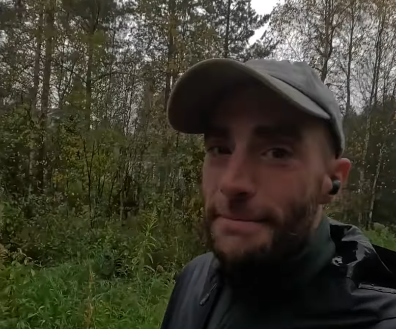 Stefano Mini sorride leggermente all'aperto, indossando un cappellino e una giacca, con un auricolare. Dietro di lui, un bosc