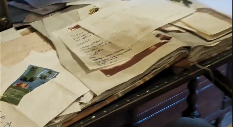 Pila di vecchie lettere e documenti su un tavolo di legno, con alcune scritte a mano visibili e una mappa parziale.