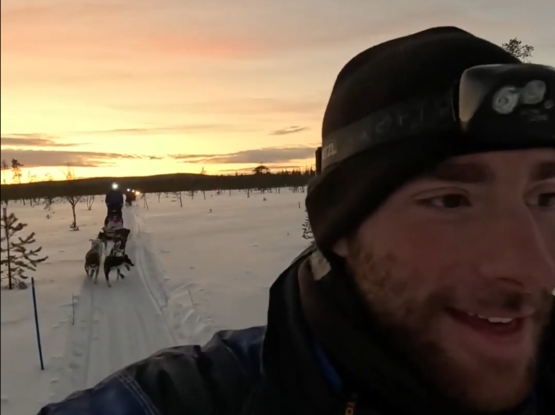 Stefano Mini sorride mentre fa dog sledding in un paesaggio innevato al tramonto, con altri slittini e cani in lontananza.