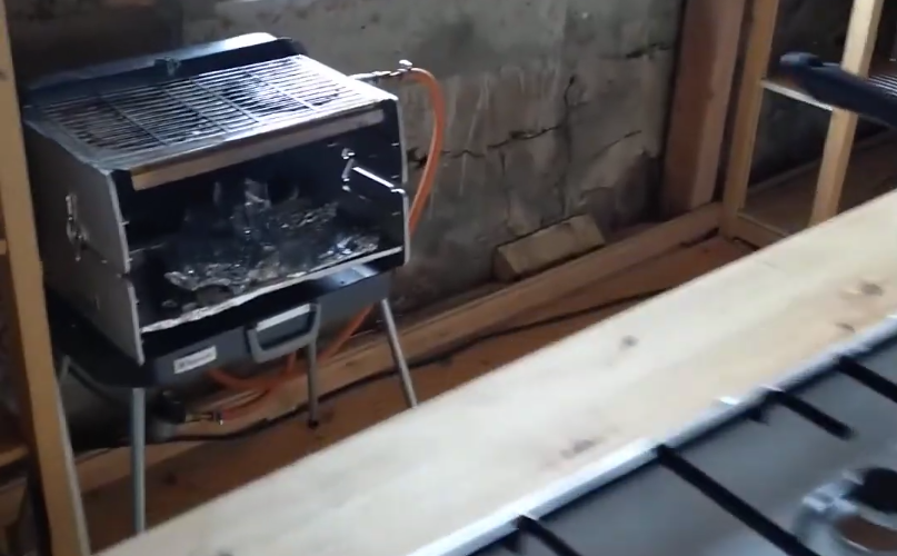 Barbecue a gas portatile con tubo di alimentazione, posizionato all'interno vicino a un muro di pietra e travi di legno.