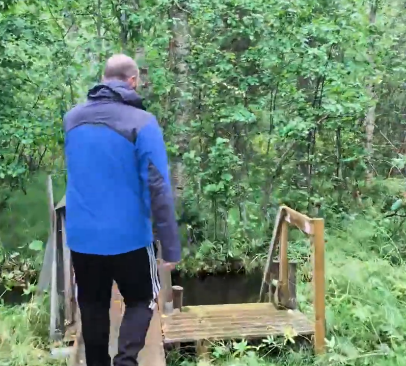 Stefano Mini cammina su un ponte di legno in una foresta lussureggiante, indossando una giacca blu e pantaloni neri.