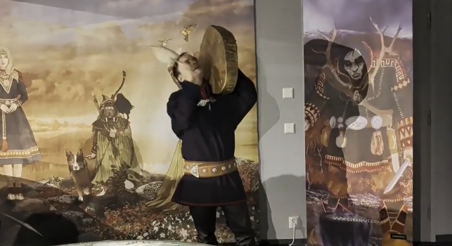 Ante Aikio in abiti tradizionali sami suona un tamburo sciamanico Sami davanti a un murale.